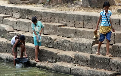 Drei Mädchen stehen auf steinernen Stufen am Wasser, eines fängt mit einem Netz Fische, ein anderes hält eine Plastikflasche, und rechts geht ein Mädchen mit einem geflochtenen Korb die Stufen hinunter.