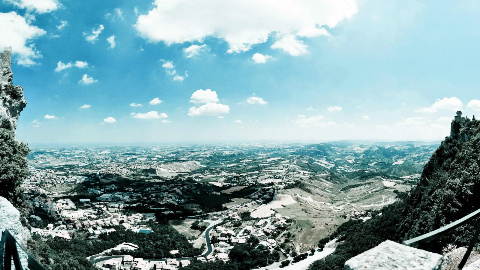 Weitblick von einer Burgmauer über Dächer, Straßen und Hügel von San Marino unter blauem Himmel mit Wolken.