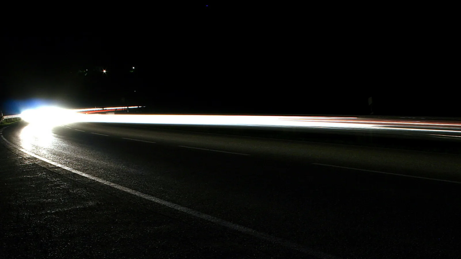 Nächtliche Straße, die sich nach links in einer Kurve verliert, mit einem hellen Scheinwerferlicht und langen roten und weißen Lichtspuren vor schwarzem Himmel.