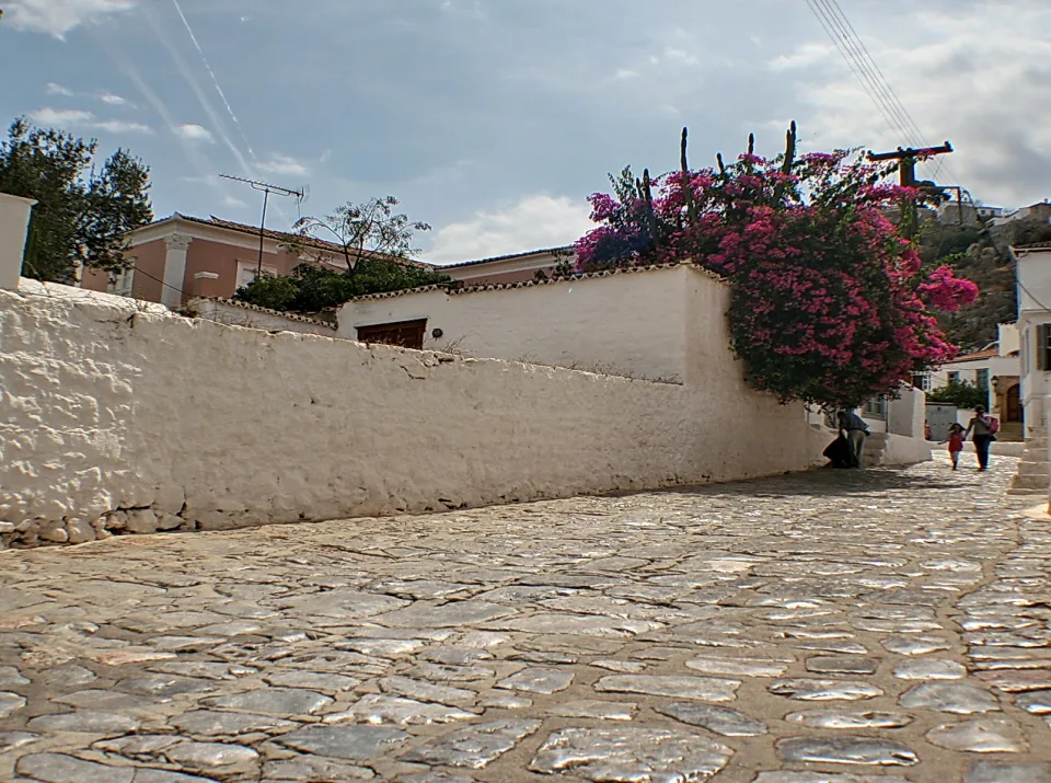 Gepflasterte Gasse zwischen weiß getünchten Mauern, über die pink blühende Bougainvillea wächst, mit zwei Passanten im Hintergrund.