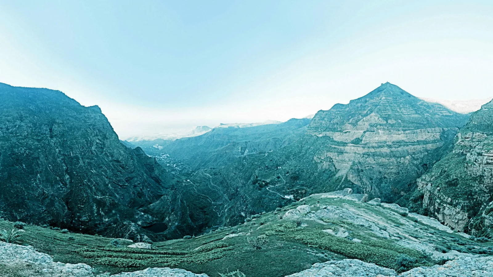 Panoramablick auf ein tief eingeschnittenes Bergtal mit grünen Terrassen im Vordergrund, steilen Felswänden und einer hellblauen, leicht diesigen Himmelsfläche.