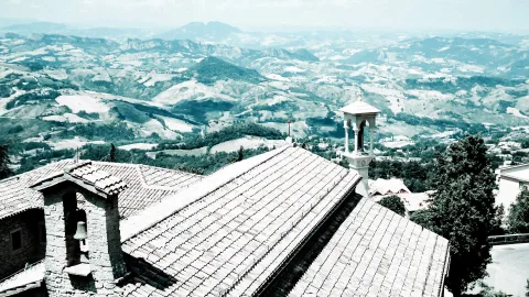 Blick von einem Dach mit Ziegeln und einem kleinen Glockenturm über eine hügelige Landschaft mit Feldern und Wäldern