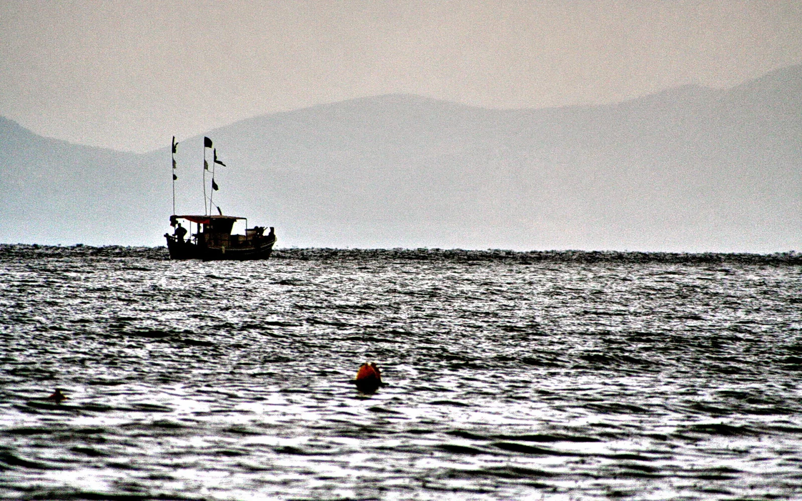Kleines Fischerboot mit Wimpeln auf unruhigem Meer vor einer dunstigen Bergkette, davor eine orange Boje im Wasser.