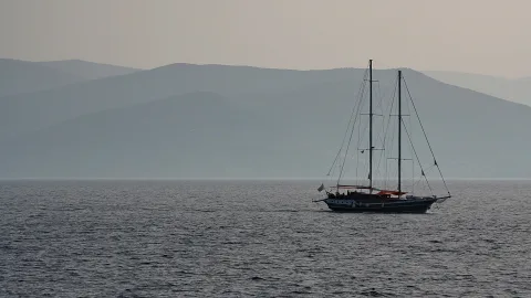 Ein zweimastiges Segelboot mit dunklem Rumpf fährt auf ruhigem, dunkelblauem Meer, im Hintergrund schichten sich bläuliche Berge im Dunst.