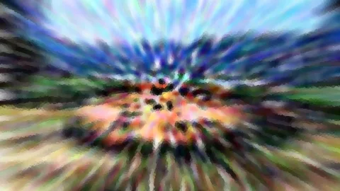 Stark verwischtes Bild mit radialem Zoom-Effekt: Im Zentrum leuchten orange und rosa Flächen mit dunklen Punkten, nach außen laufen blaue, grüne und gelbe Schlieren, oben ein helles Himmelsblau.