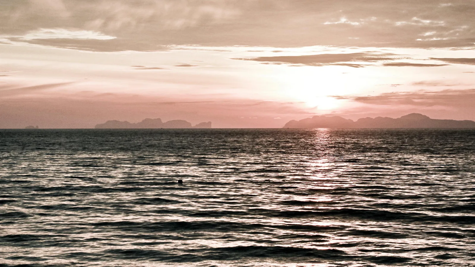 Sonnenuntergang über einer ruhigen Meeresoberfläche, im Hintergrund dunkle Inselsilhouetten unter rosa getönten Wolken