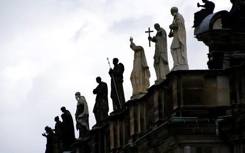 Eine Reihe von Heiligenstatuen aus Stein steht auf einer Dachbrüstung vor bewölktem Himmel, einige Figuren tragen Kreuz, Stab oder Gewänder.