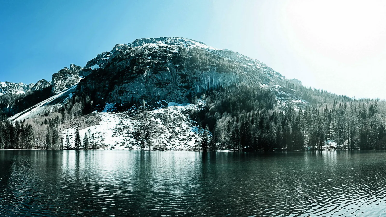 Breites Panorama eines Bergsees mit leicht gekräuselter Wasseroberfläche. Dahinter steigt eine felsige Bergflanke auf, deren Gipfel und Hänge teilweise mit Schnee bedeckt sind. Am Ufer stehen dunkle Nadelbäume, der Himmel ist klar und blau.