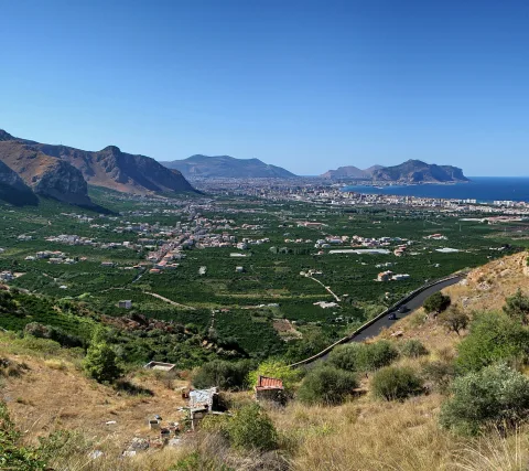 Weiter Blick von einem bewachsenen Hügel auf eine grüne Ebene mit Häusern, eine Stadt am Meer und zwei Bergrücken unter klarem blauem Himmel.