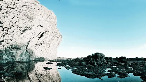 Helle Kalkfelsklippe links, deren Spiegelung im flachen Wasser liegt, rechts dunkle Felsen zwischen Tümpeln unter blauem Himmel.