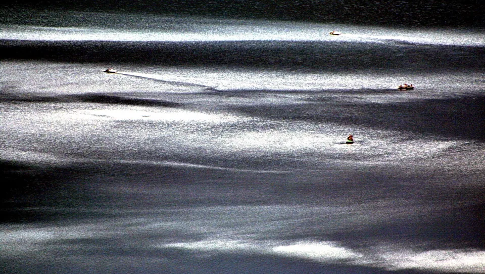 Mehrere kleine Boote mit Personen auf einer silbrig schimmernden Wasseroberfläche mit dunklen Streifen.