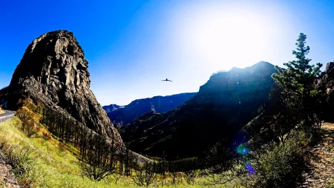 Ein hoher, dunkler Vulkanfelsen links neben einer Straße, dahinter ein tiefes Tal mit Bergrücken unter blauem Himmel und strahlender Sonne, ein Vogel fliegt am Himmel.