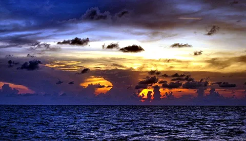 Dramatische Wolkenformation über dem Meer, die Sonne ist kurz über dem Horizont und färbt den Himmel orange und gelb, darunter dunkles, leicht bewegtes Wasser.