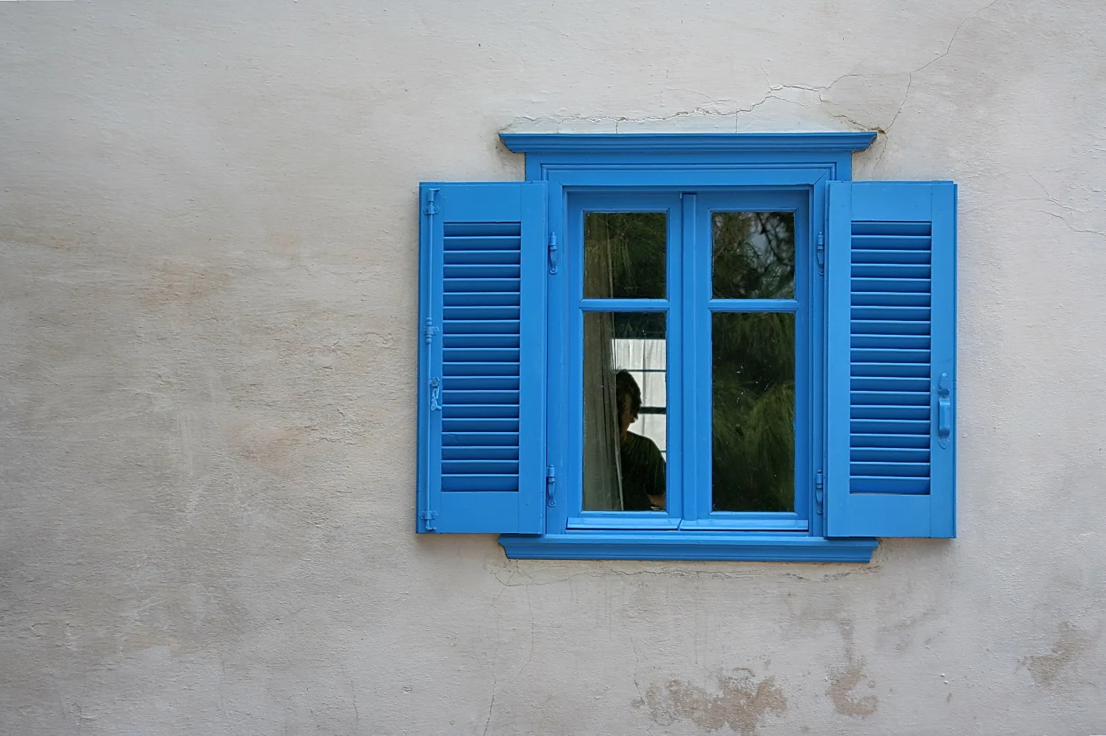 Blau gestrichenes Fenster mit geöffneten Fensterläden in einer hellen, verputzten Wand; hinter der Scheibe ist eine Person als Silhouette zu erahnen.