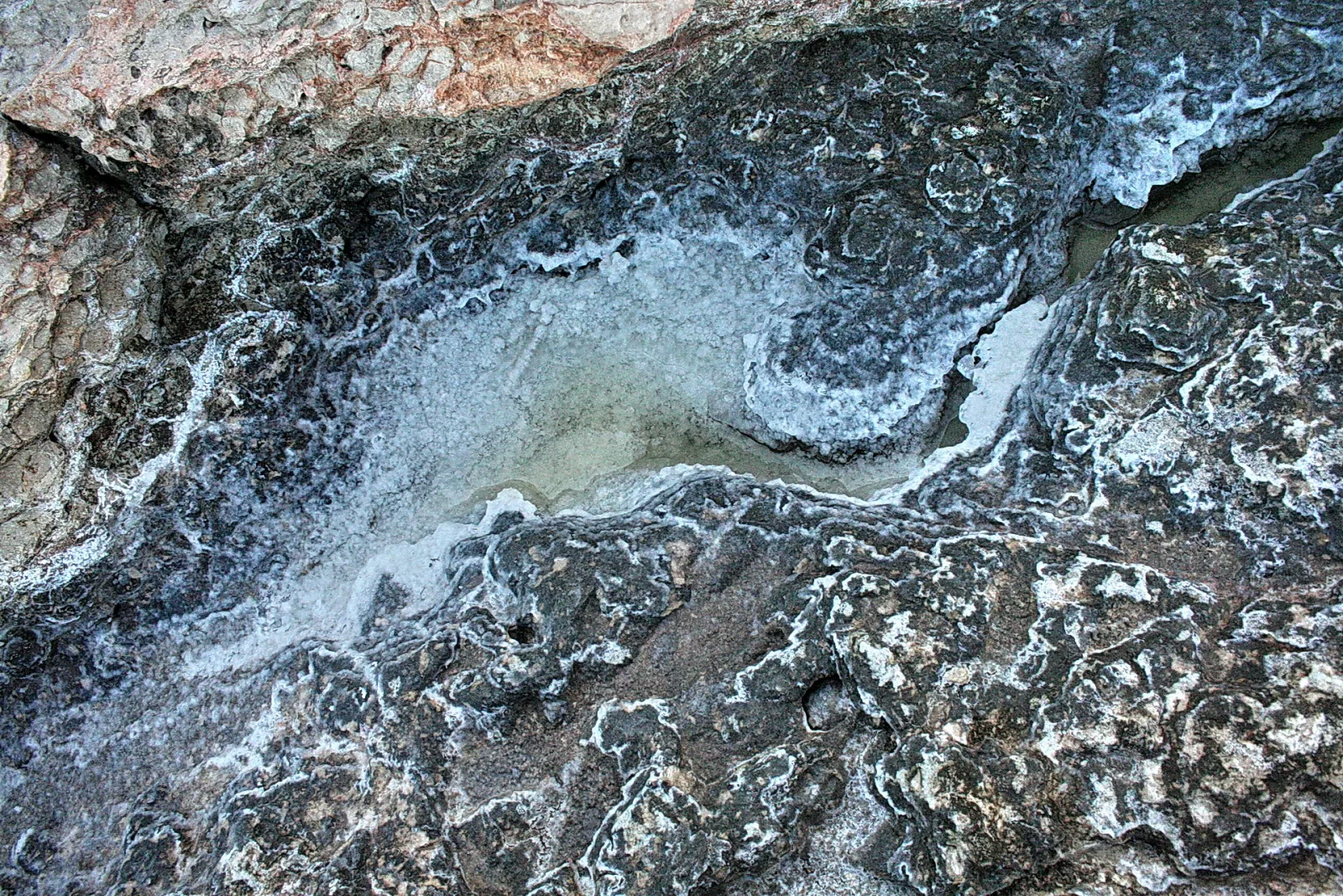 Graublaue Felsen am Meer mit weißen Salzkrusten um eine flache, ausgetrocknete Pfütze.