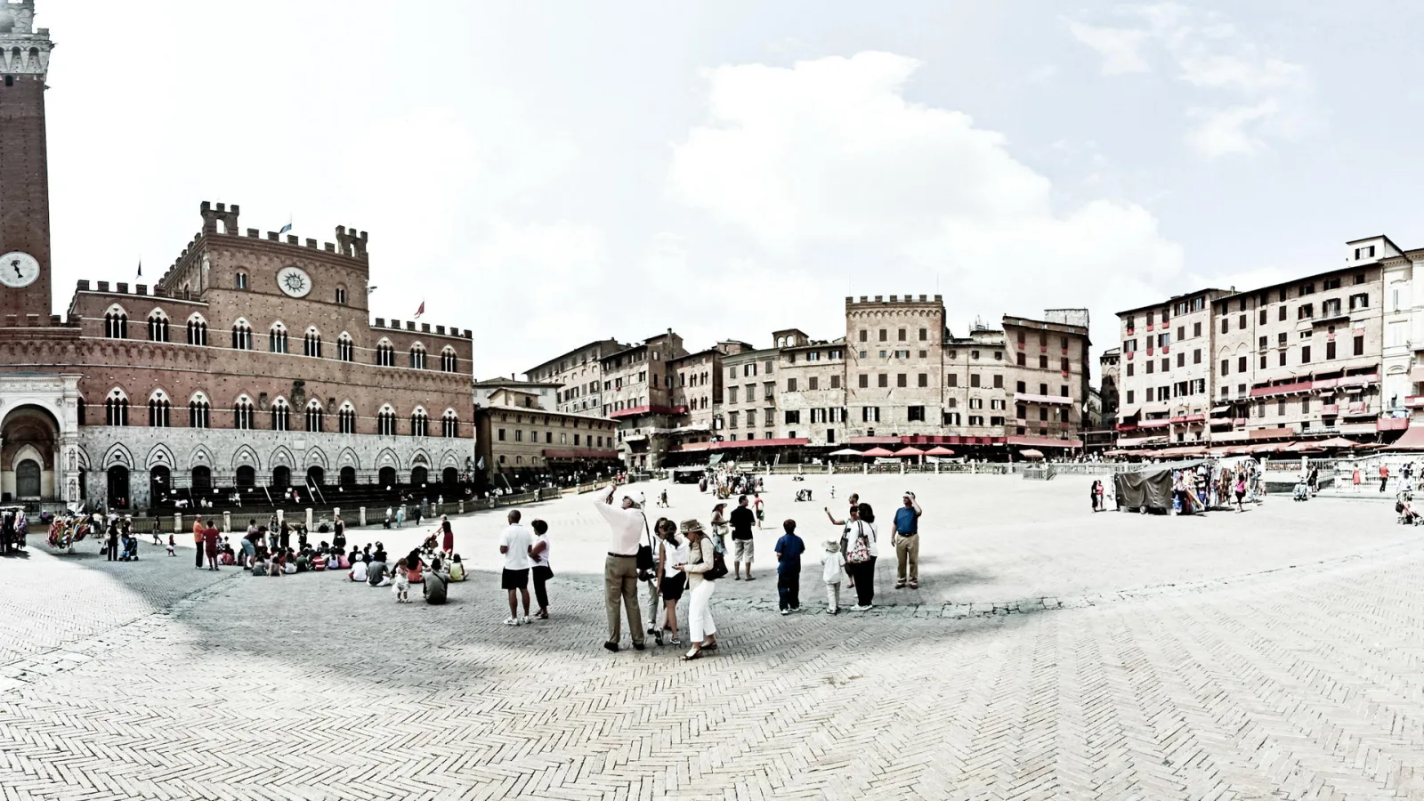 Belebter Marktplatz aus Kopfsteinpflaster in Siena, im Vordergrund stehen und sitzen Touristen, links ragt ein mittelalterlicher Backsteinpalast mit Uhr und Turm auf, rechts reihen sich Häuser mit roten Sonnenschirmen unter blauem Himmel.