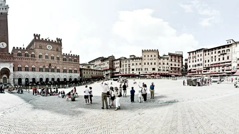 Belebter Marktplatz aus Kopfsteinpflaster in Siena, im Vordergrund stehen und sitzen Touristen, links ragt ein mittelalterlicher Backsteinpalast mit Uhr und Turm auf, rechts reihen sich Häuser mit roten Sonnenschirmen unter blauem Himmel.