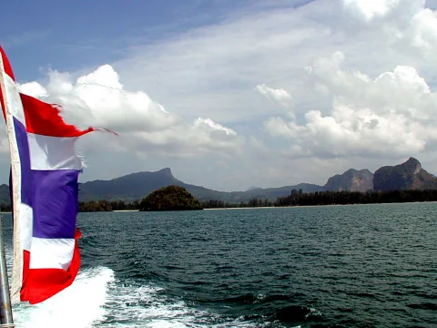 Eine thailändische Flagge weht am Heck eines Bootes, dahinter liegt ruhiges Meer mit bewaldeten Inseln und felsigen Bergen unter einem Himmel mit Wolken.