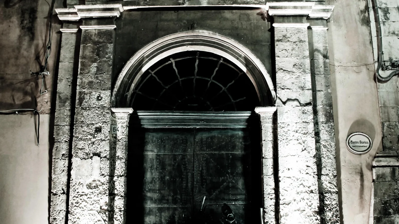 Schwarzweiß-Foto eines Barockportals mit halbrundem Oberlicht und Gitterfenster, flankiert von steinernen Pilastern, nachts an einer alten Hausfassade in Noto, Sizilien.