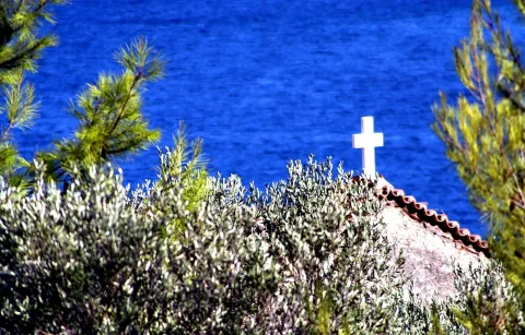 Weißes Kreuz auf einem Ziegeldach zwischen Olivenbäumen und Kiefern, dahinter das blaue Meer.