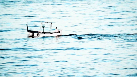 Ein weißes Fischerboot mit schwarzem Rand und zwei Rettungsringen fährt von links nach rechts über eine leicht gekräuselte blaugraue See. Eine einzelne Person sitzt am Heck.