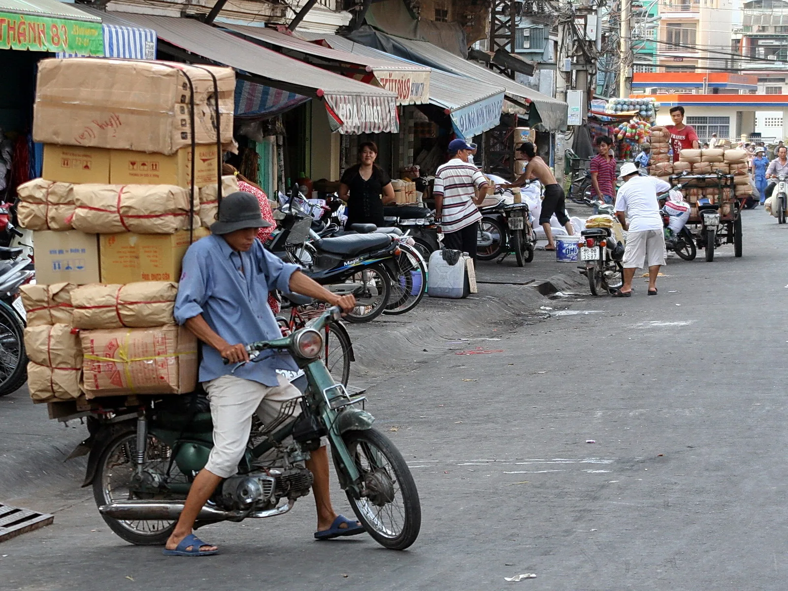 Ein Mann auf einem grünen Mofa transportiert hoch gestapelte Kartons mit Ware durch eine belebte Straße, im Hintergrund Marktstände, Passanten und weitere Motorroller.