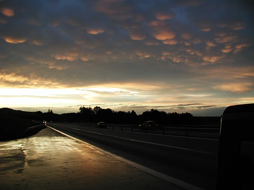 Nasse Autobahn im Abendlicht, darüber dunkle Wolken mit orangefarbenen Unterseiten, rechts eine Leitplanke und dunkle Bäume am Horizont.