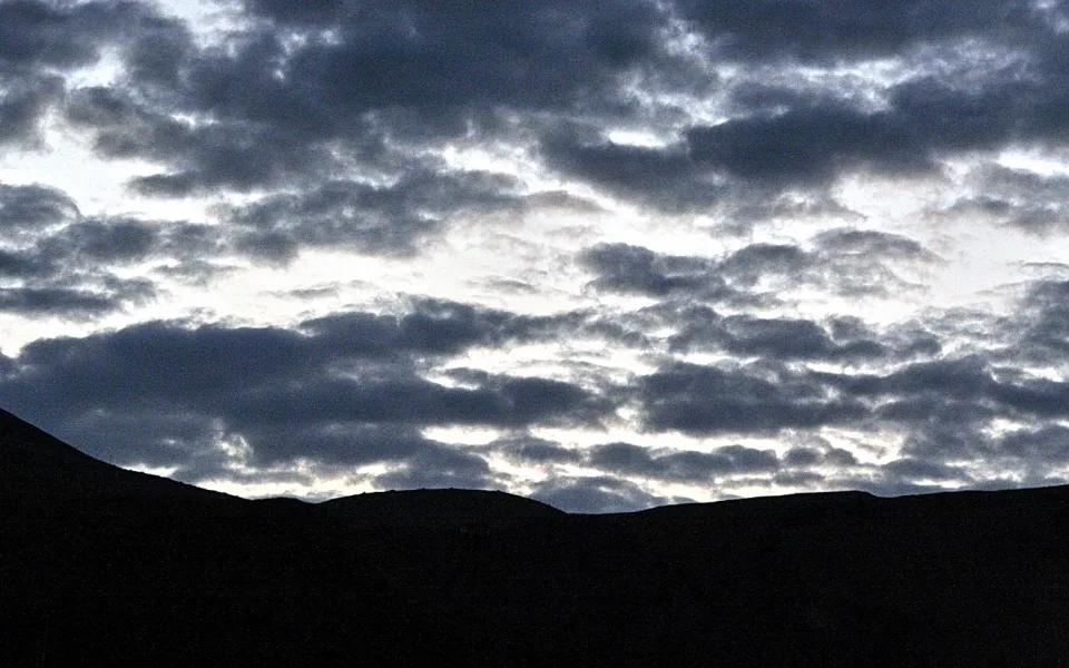 Dunkle Hügelkette als Silhouette vor einem bewölkten Abendhimmel mit hellen Lücken zwischen den Wolken