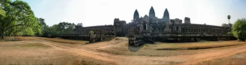 Panoramaaufnahme des Tempels Angkor Wat mit mehreren Türmen hinter einer Mauer, davor eine staubige Erdstraße und Bäume links