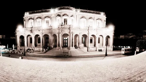 Beleuchtetes barockes Gebäude mit Bogengängen und Balkonbrüstung auf einem Platz in Noto bei Nacht, in Sepiatönen, davor eine breite Treppe.