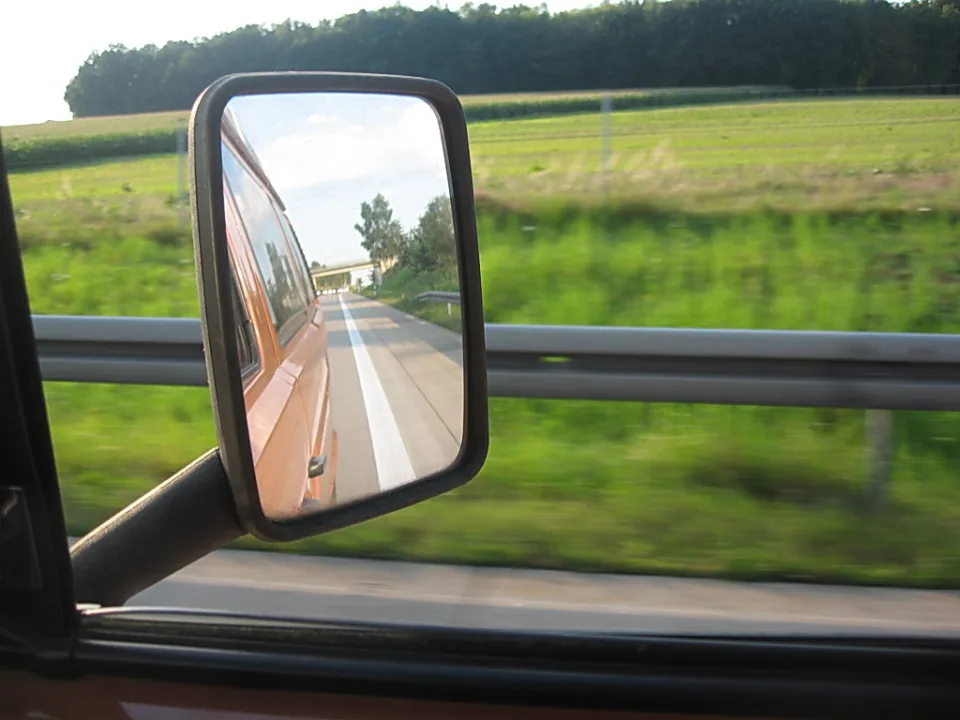 Blick durch ein Autofenster auf einen Seitenspiegel, der eine asphaltierte Landstraße mit weißer Mittellinie, einen orangefarbenen Wagen und Bäume zeigt; davor eine Leitplanke und unscharfe grüne Felder.