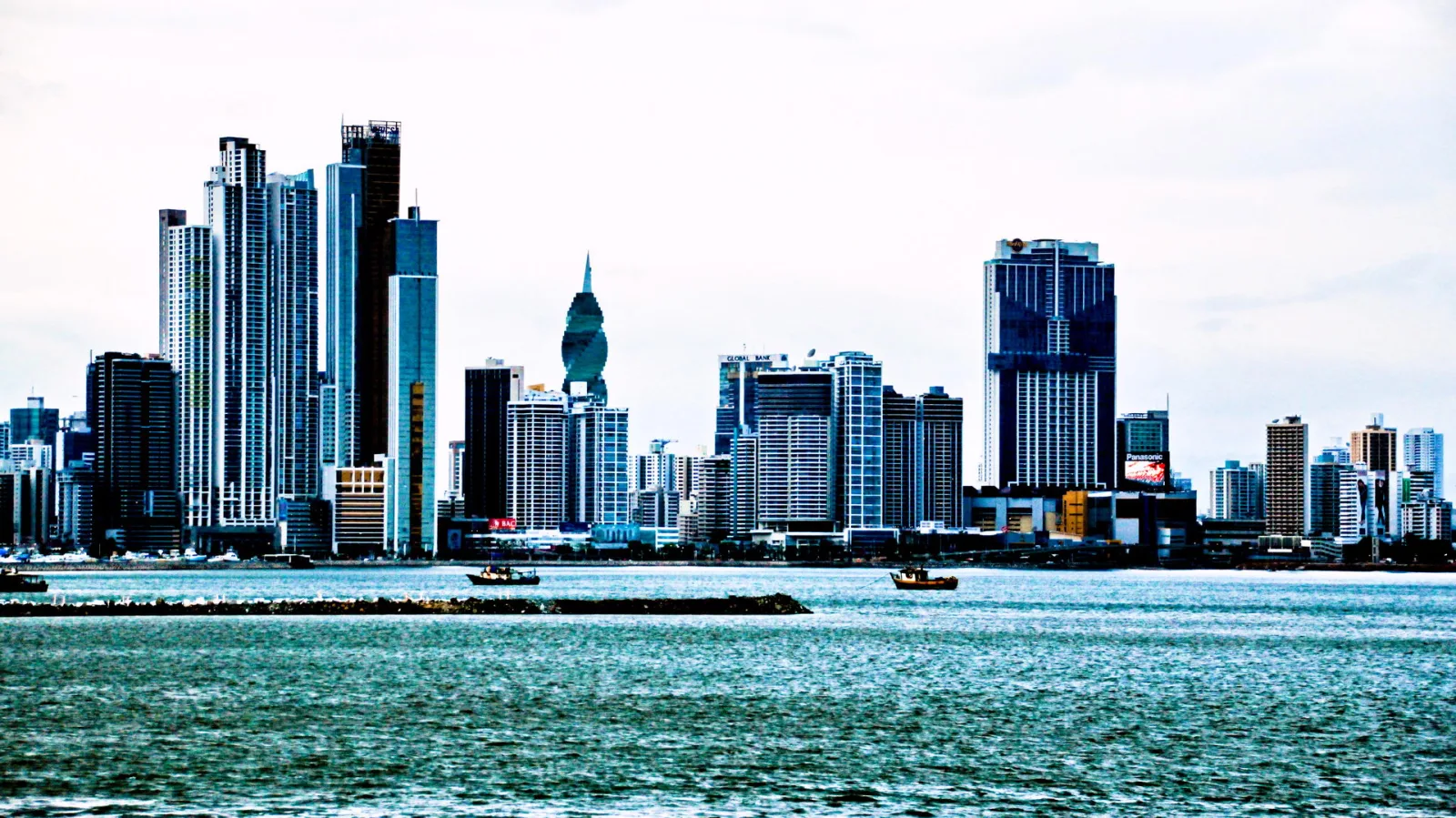 Dichte Skyline mit Hochhäusern am Wasser unter bewölktem Himmel, davor zwei kleine Boote und ein langer Damm.