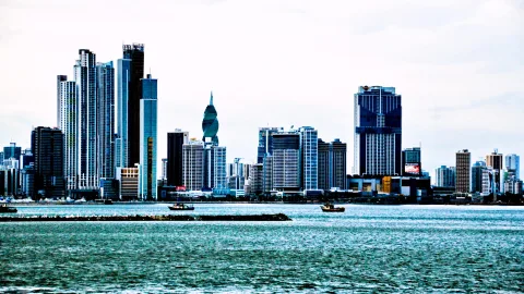 Dichte Skyline mit Hochhäusern am Wasser unter bewölktem Himmel, davor zwei kleine Boote und ein langer Damm.