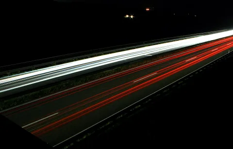 Langzeitaufnahme einer nächtlichen Autobahn mit weißen Scheinwerferspuren auf der linken Fahrbahn und roten Rücklichtspuren auf der rechten, im Hintergrund zwei Lichter am dunklen Horizont.