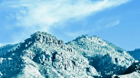 Zwei bewaldete Bergkuppen mit steilen Felswänden unter blauem Himmel mit Wolkenschleiern.