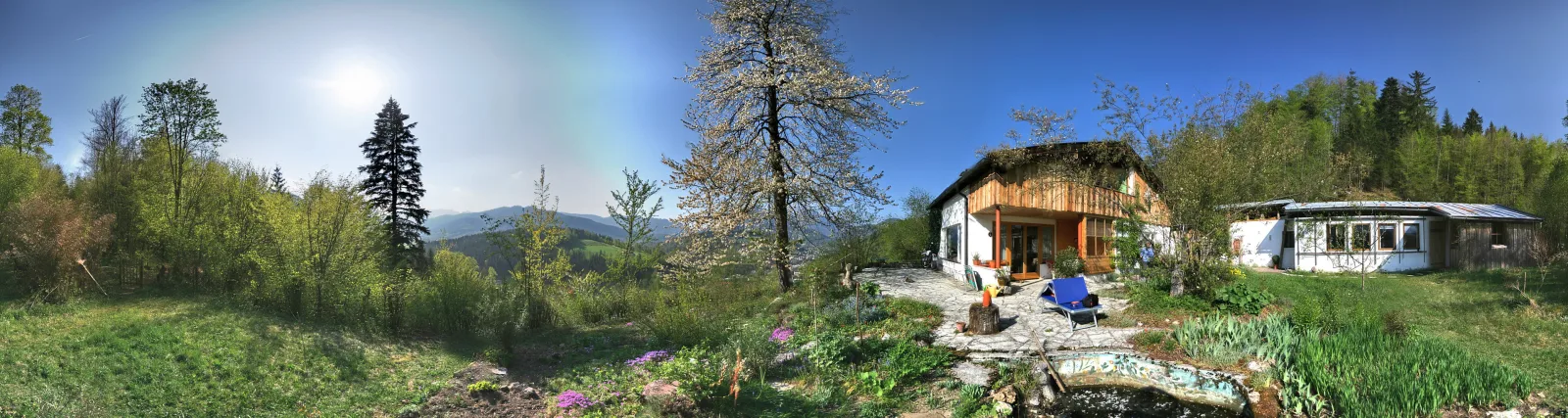 Panoramablick auf ein Holzhaus mit Terrasse und blauem Stuhl, davor ein blühender Baum und Blumenbeete, dahinter bewaldete Hügel unter blauem Himmel mit Sonne.