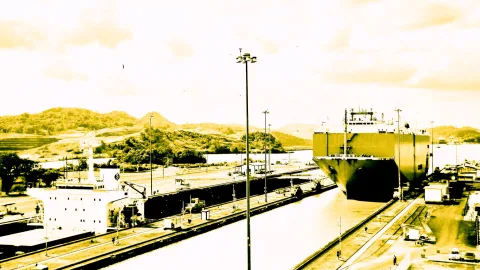 Großes Frachtschiff liegt in einer Schleusenkammer des Panama-Kanals, links ein weiteres Schiff, dahinter bewaldete Hügel unter einem hellen Himmel.