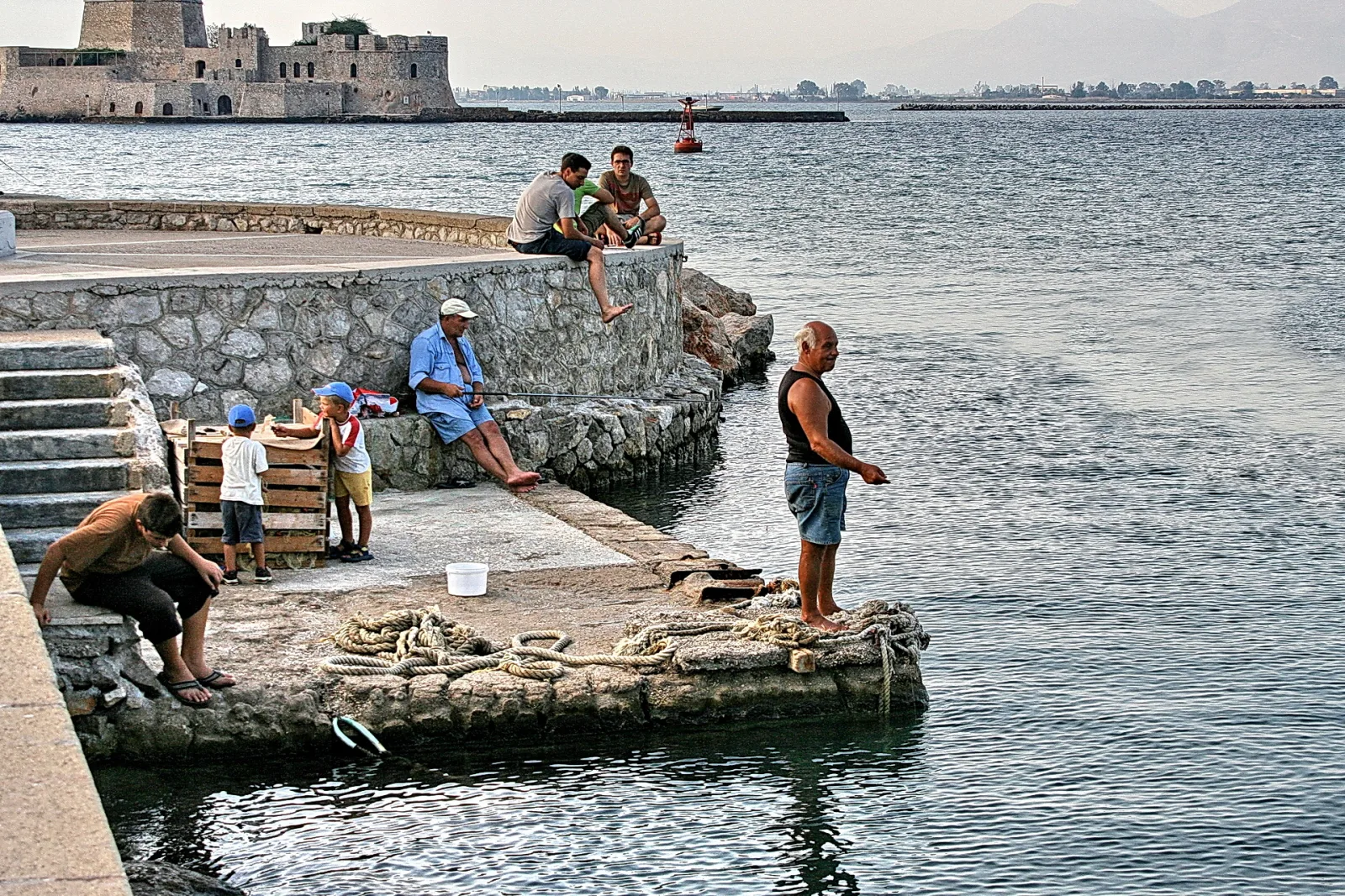 Mehrere Männer und zwei Jungen sitzen und stehen auf einer Steinmole am Wasser, im Hintergrund eine alte Festung am Meer.