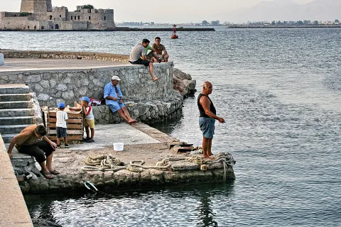 Mehrere Männer und zwei Jungen sitzen und stehen auf einer Steinmole am Wasser, im Hintergrund eine alte Festung am Meer.