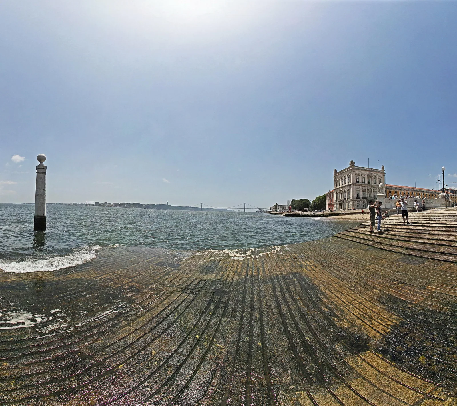 Breite, flach ins Wasser abfallende Steintreppe am Tejo, links eine weiße Säule mit Kugel, rechts ein historisches Gebäude mit Menschen auf der Uferpromenade, im Hintergrund eine Hängebrücke unter hellem Himmel.