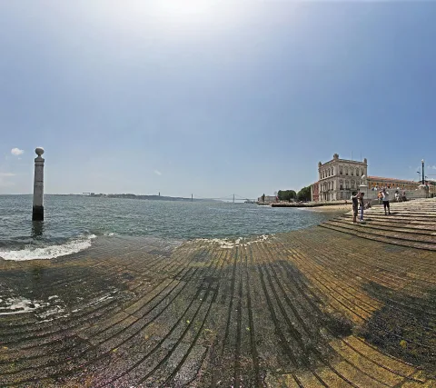 Breite, flach ins Wasser abfallende Steintreppe am Tejo, links eine weiße Säule mit Kugel, rechts ein historisches Gebäude mit Menschen auf der Uferpromenade, im Hintergrund eine Hängebrücke unter hellem Himmel.