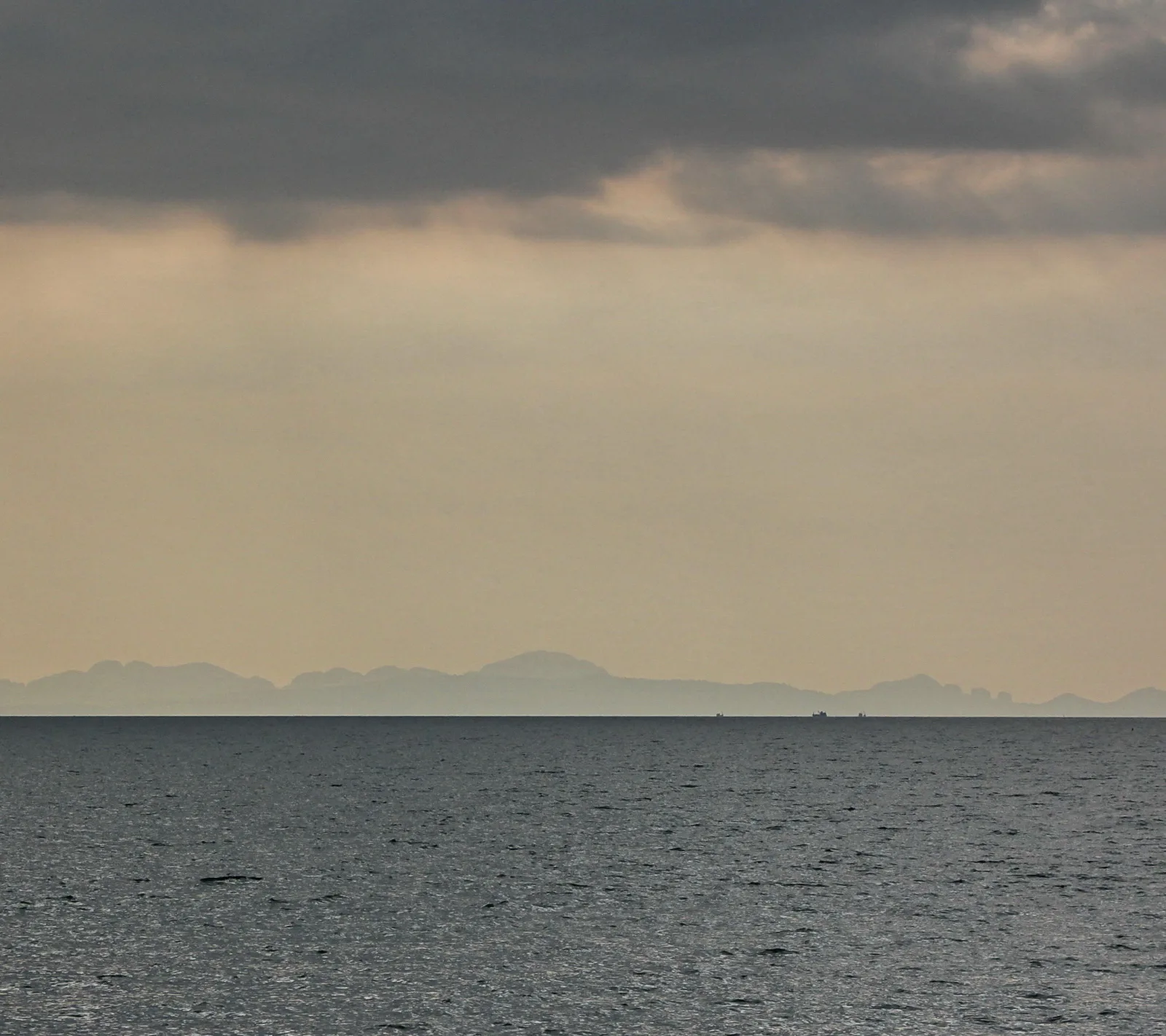 Ruhige graue See unter dichten Wolken, am Horizont eine blasse Bergkette und zwei winzige Schiffe.