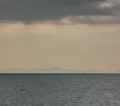 Ruhige graue See unter dichten Wolken, am Horizont eine blasse Bergkette und zwei winzige Schiffe.