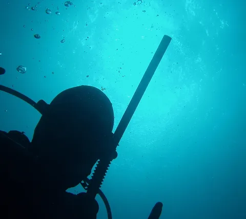 Silhouette eines Tauchers mit Atemregler vor blauem Wasser, aus dem Luftblasen nach oben steigen