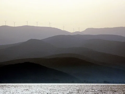 Mehrere Hügelketten staffeln sich in dunstigem Licht bis zum Horizont, auf dem Kamm der hinteren Kette stehen sieben Windräder. Im Vordergrund liegt glitzerndes Wasser.