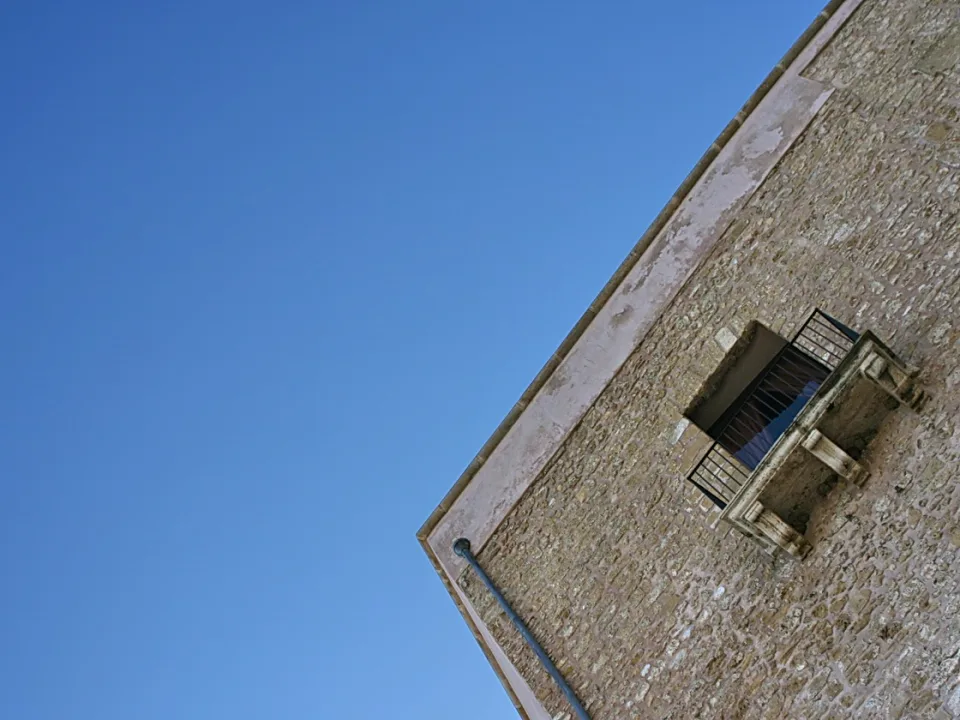 Ecke eines Steinhauses mit vergittertem Fenster und Balkon vor klar blauem Himmel.