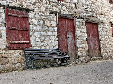 Alte Steinmauer mit roten, verwitterten Holztüren und Fensterläden, davor eine schwarze Gusseisenbank auf einem Schotterweg.