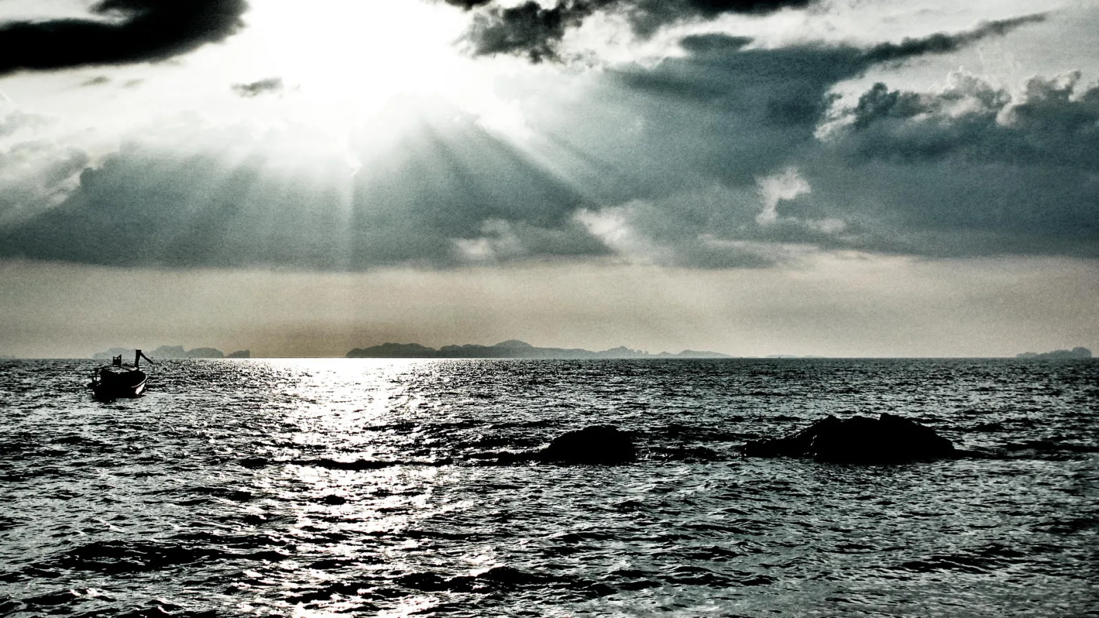 Sonnenstrahlen brechen durch dunkle Wolken über einem Meer, links ein kleines Boot, im Vordergrund zwei Felsen im Wasser, am Horizont Inseln.