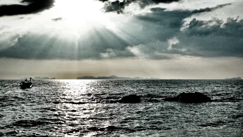 Sonnenstrahlen brechen durch dunkle Wolken über einem Meer, links ein kleines Boot, im Vordergrund zwei Felsen im Wasser, am Horizont Inseln.