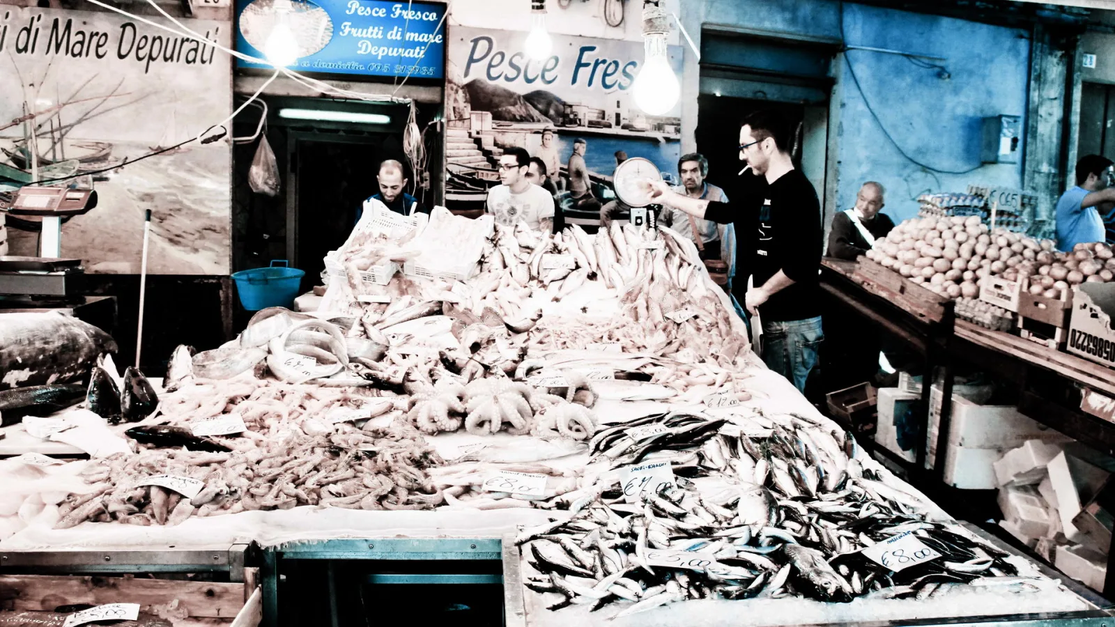 Fischstand auf einem Markt mit Bergen von Tintenfischen, Garnelen und Sardinen auf Eis, dahinter Verkäufer und Kisten mit Kartoffeln.